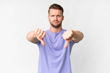 İzole edilmiş beyaz arka planda genç, yakışıklı, beyaz bir adam baş parmağını iki eliyle aşağı gösteriyor.