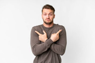 Young handsome caucasian man over isolated white background pointing to the laterals having doubts