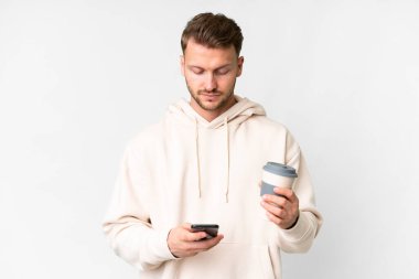 İzole edilmiş beyaz arka planı olan yakışıklı beyaz bir adam elinde kahve ve bir cep telefonu tutuyor.