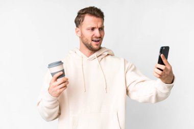 İzole edilmiş beyaz arka planı olan yakışıklı beyaz bir adam elinde kahve ve bir cep telefonu tutuyor.
