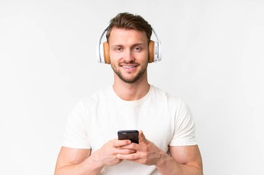 İzole edilmiş beyaz arka plan üzerinde yakışıklı beyaz bir adam cep telefonuyla müzik dinliyor ve ön tarafa bakıyor.