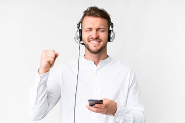 Tele pazarlamacı beyaz kulaklıklı bir adam zafer pozisyonunda telefon ile izole edilmiş beyaz arka planda çalışıyor.
