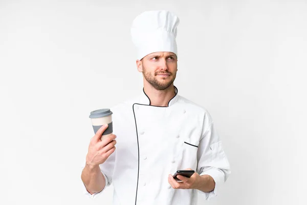 年轻的高加索厨师站在孤立的白色背景之上 一边拿着咖啡 一边想着什么 一边拿着手机 — 图库照片