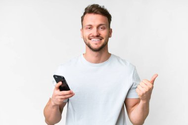 İzole edilmiş beyaz arka plan üzerinde genç, yakışıklı beyaz adam başparmak kaldırırken cep telefonu kullanıyor.