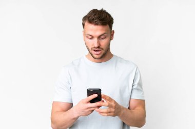 Beyaz arka plan üzerinde genç, yakışıklı beyaz bir adam kameraya bakarken şaşırmış bir ifadeyle cep telefonunu kullanıyor.