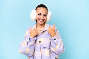 Mavi arka planda izole edilmiş kışlık kuklalar takan genç bir Arap kadın. Baş parmakları havada ve gülümsüyor.