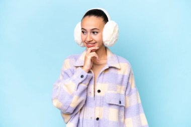 Mavi arka planda izole edilmiş kışlık kuklalar giyen genç bir Arap kadın yan tarafa bakıyor.