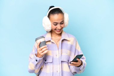Mavi arka planda izole edilmiş kışlık kuklalar takan genç bir Arap kadın elinde kahve ve bir cep telefonu tutuyor.