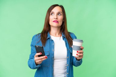 Orta yaşlı bir kadın izole edilmiş krom anahtar arka planda elinde kahve ve bir şeyler düşünürken bir cep telefonu tutuyor.