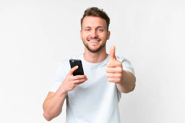 年轻英俊的白人男子在竖起大拇指的时候用手机在孤立的白人背景上 — 图库照片