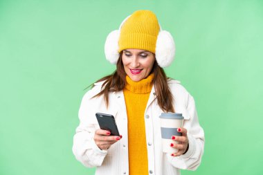 Orta yaşlı bir kadın, izole edilmiş krom arka planda kış maskesi takıyor. Elinde kahve ve cep telefonu var.