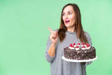 Orta yaşlı beyaz kadın izole edilmiş krom anahtar arkaplan üzerinde doğum günü pastası tutuyor. Çözümü bulmak için parmağını kaldırıyor.