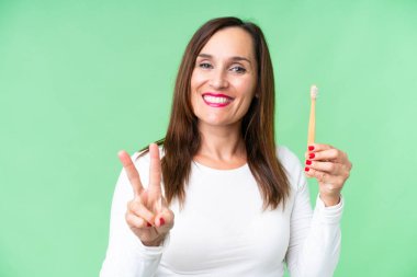 Orta yaşlı bir kadın izole edilmiş krom arka planda dişlerini fırçalıyor gülümsüyor ve zafer işareti gösteriyor.