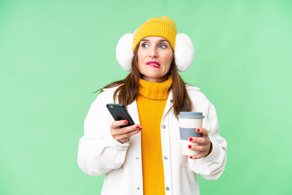 Женщина средних лет носит зимние муфты на изолированном хрома ключ фоне держа кофе, чтобы забрать и мобильный, думая что-то