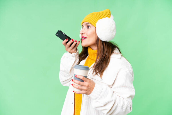 Женщина средних лет носит зимние муфты на изолированном хрома ключ фоне держа кофе, чтобы забрать и мобильный