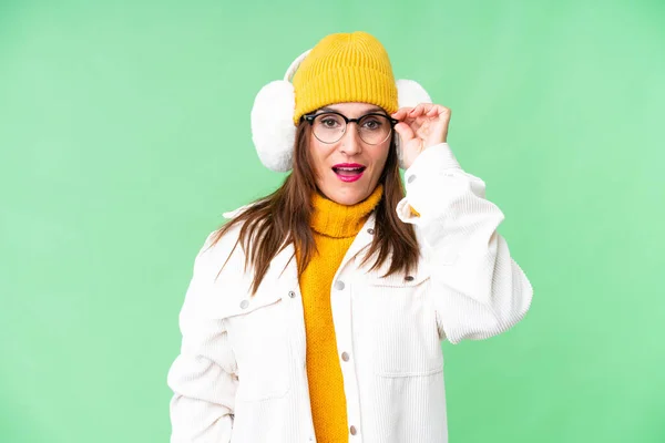 stock image Middle age woman wearing winter muffs over isolated chroma key background with glasses and surprised