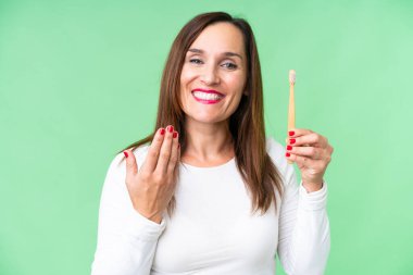 Orta yaşlı bir kadın, izole edilmiş krom arka planda dişlerini fırçalıyor ve elini uzatmaya davet ediyor. Gelmene sevindim.