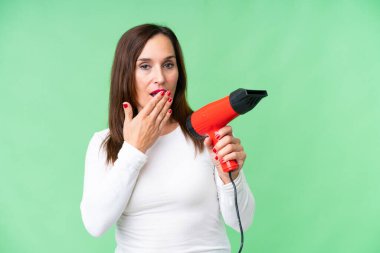 Orta yaşlı bir kadın elinde saç kurutma makinesiyle izole edilmiş krom anahtar arka planda şaşırtıcı ve şok olmuş bir yüz ifadesiyle duruyor.