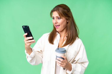 Orta yaşlı bir kadın izole edilmiş arka planda kahve ve bir cep telefonu tutuyor.