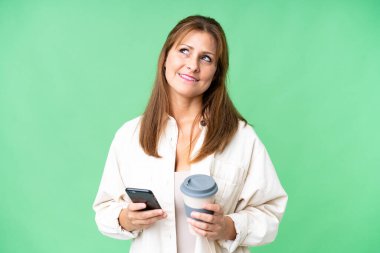 Orta yaşlı bir kadın, izole edilmiş arka planda kahve ve bir şeyler düşünürken cep telefonu tutuyor.