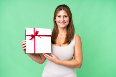 Orta yaşlı, beyaz bir kadın, izole edilmiş arka planda elinde bir hediye tutuyor. Çok gülümsüyor.