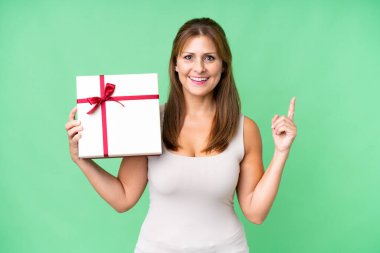 Orta yaşlı beyaz bir kadın, izole bir arka plan üzerinde elinde bir hediye tutuyor. Harika bir fikre işaret ediyor.