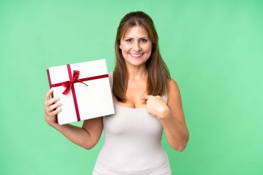 Orta yaşlı, beyaz bir kadın, izole edilmiş bir geçmişe sahip yüzünde sürpriz bir ifadeyle bir hediye taşıyor.