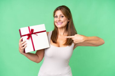 Orta yaşlı beyaz bir kadın, izole edilmiş arka planda bir hediye tutuyor ve onu işaret ediyor.