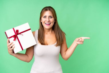 Orta yaşlı, beyaz bir kadın, izole bir arka planda elinde bir hediye tutuyor. Şaşırmış ve parmağıyla onu işaret ediyor.