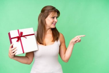 Orta yaşlı beyaz bir kadın, izole edilmiş arka plan üzerinde bir hediye tutuyor ve bir ürün sunmak için bir tarafa bakıyor.