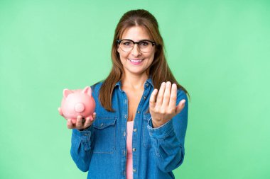 Orta yaşlı, beyaz bir kadın, izole bir arka planda kumbarayı elinde tutuyor ve elini kolunu sallayarak gelmeye davet ediyor. Gelmene sevindim.