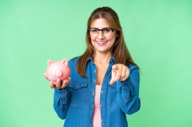 Orta yaşlı, beyaz bir kadın izole bir arka planda kumbarayı elinde tutuyor. Kendine güvenen bir ifadeyle seni işaret ediyor.