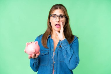 Orta yaşlı beyaz bir kadın izole bir arka planda domuz kumbarası tutuyor ve ağzı açık bağırıyor.