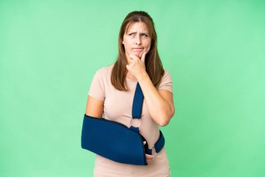 Middle age with broken arm and wearing a sling over isolated background having doubts and thinking