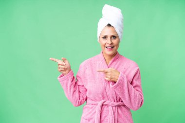 Soyutlanmış arka planda, beyaz bornoz giymiş orta yaşlı bir kadın şaşırmış ve onu işaret ediyor.