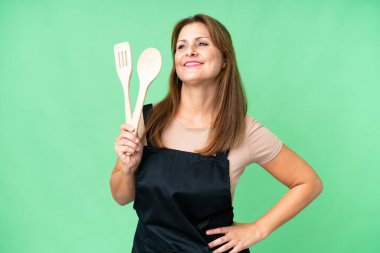 Orta yaşlı, beyaz bir kadın, izole edilmiş bir arka planda, kolları kalçasında ve gülümseyerek poz veren bir rozet tutuyor.