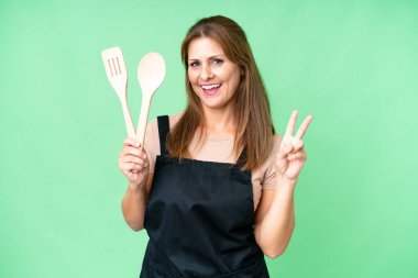 Orta yaşlı, beyaz bir kadın, izole edilmiş arka planda bir rozet tutuyor. Gülümsüyor ve zafer işareti gösteriyor.