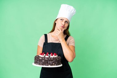 Orta yaşlı pasta şefi kadın izole edilmiş arka planda büyük bir pasta tutuyor. Şüpheleri var.