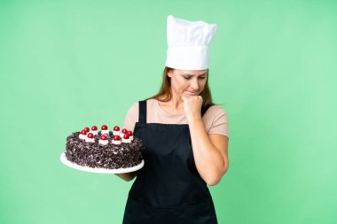 Orta yaşlı pasta şefi kadın izole edilmiş arka planda büyük bir pasta tutuyor. Şüpheleri var.