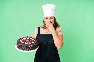 Orta yaşlı pasta şefi kadın izole edilmiş arka planda büyük bir pasta tutuyor mutlu ve ağzı kapalı gülümsüyor.