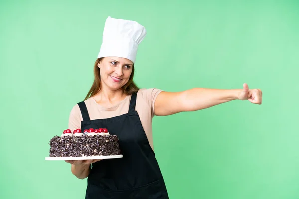 Middle age pastry chef woman holding a big cake over isolated background giving a thumbs up gesture