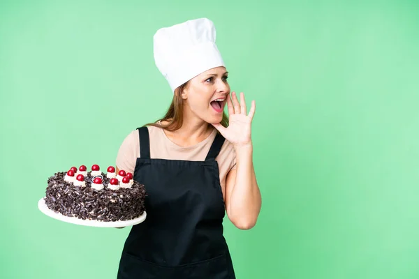 Middle age pastry chef woman holding a big cake over isolated background shouting with mouth wide open to the side