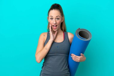 Genç beyaz kadın yoga derslerine giderken elinde mavi arka planda izole edilmiş bir minder tutarken şaşırmış ve şok olmuş bir yüz ifadesi vardı.