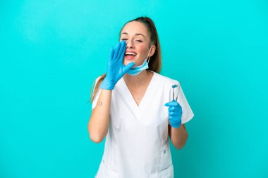 Dişçi beyaz kadın, elinde mavi arka planda izole edilmiş aletlerle ağzı açık bağırıyor.