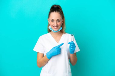 Beyaz dişçi kadının elinde mavi arka planda izole edilmiş aletler var. Bir ürünü sunmak için yan tarafı işaret ediyor.