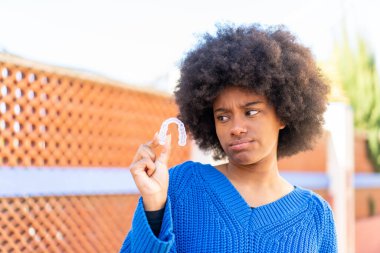 Afro-Amerikalı kız, dışarıda, üzgün bir ifadeyle görünmez diş telleri tutuyor.