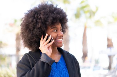 Afro-Amerikalı kız dışarıda cep telefonuyla birileriyle sohbet ediyor.