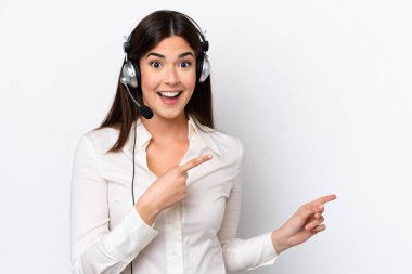 Beyaz arka planda izole edilmiş kulaklıkla çalışan beyaz pazarlamacı kadın şaşırmış ve işaret ediyor.