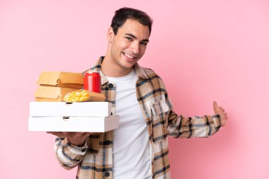 Young caucasian man holding fast food isolated on pink background extending hands to the side for inviting to come