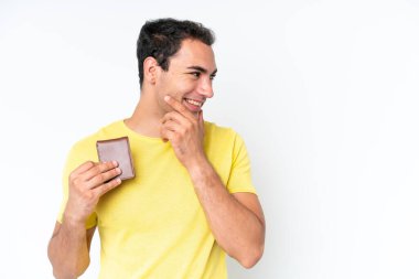 Beyaz arka planda bir cüzdan tutan genç beyaz bir adam bir fikir düşünüyor ve bakıyor.